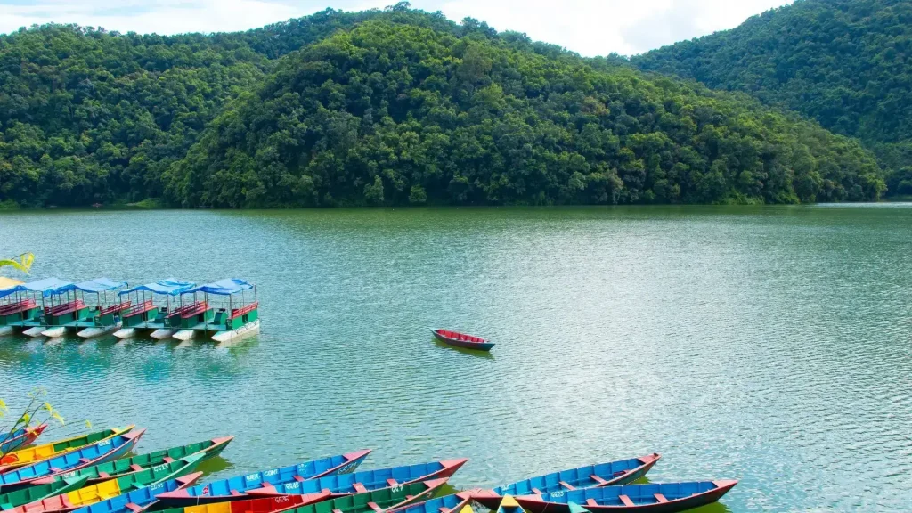 Rowboats in blue and orange float on the calm surface of Pokhara Lake, surrounded by mountains.