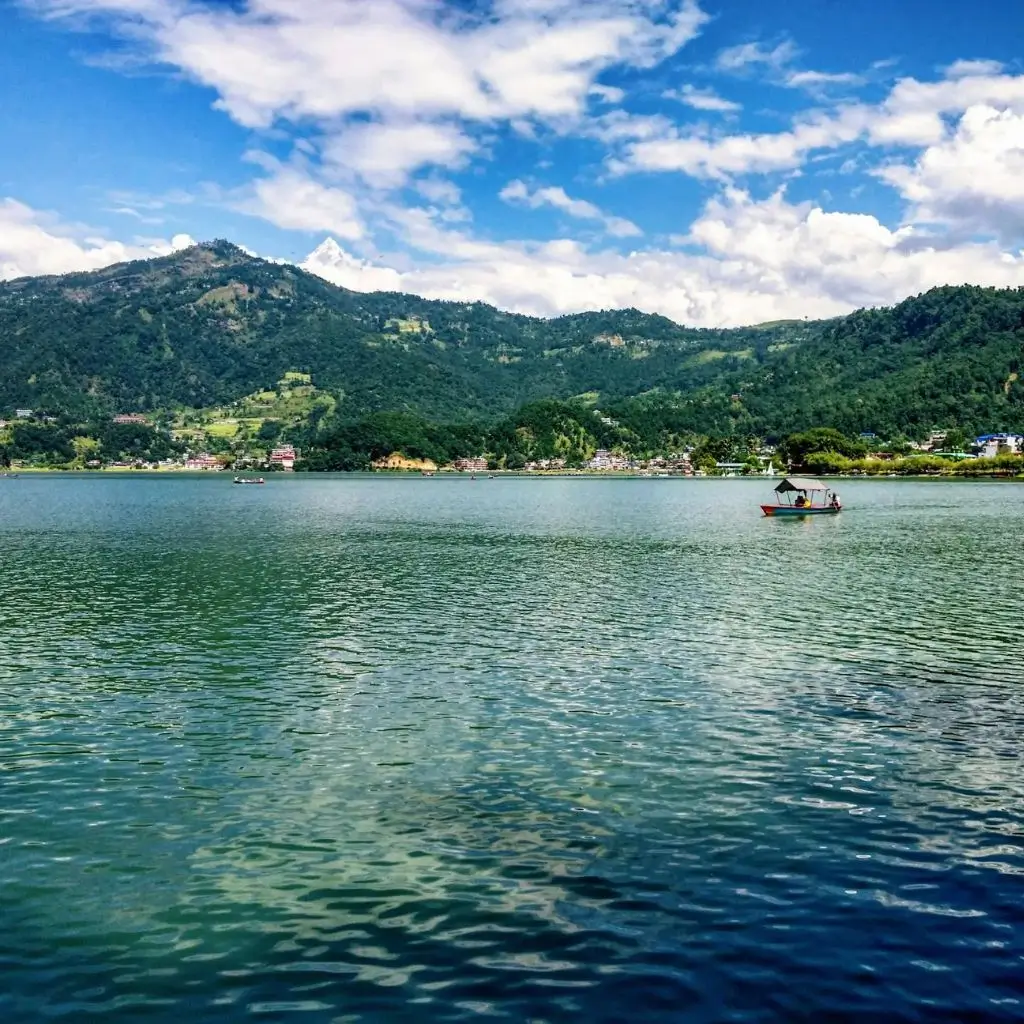 Rowboats in blue and orange float on the calm surface of Pokhara Lake, surrounded by mountains.